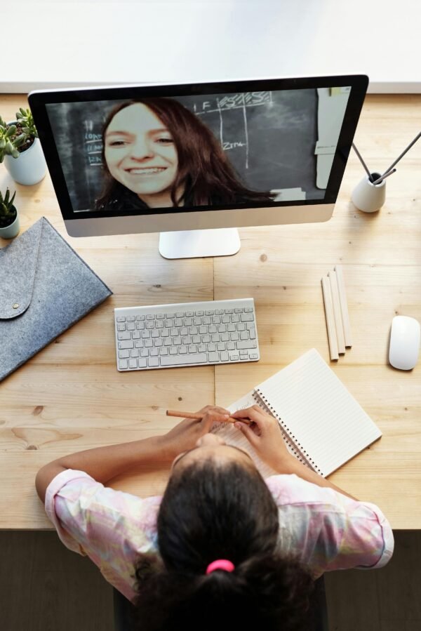 Teen girl participates in online class from home, focused on a computer screen with notebook.